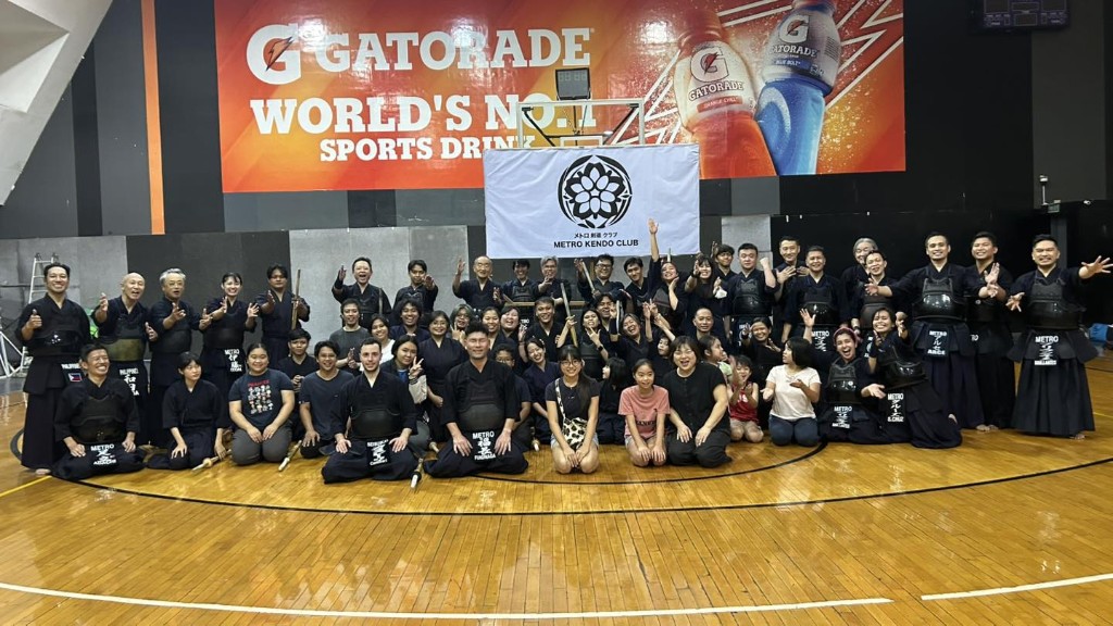 Metro Kendo Club members in kendo uniforms and armor pose for a group photo in a gymnasium, with the club banner behind them